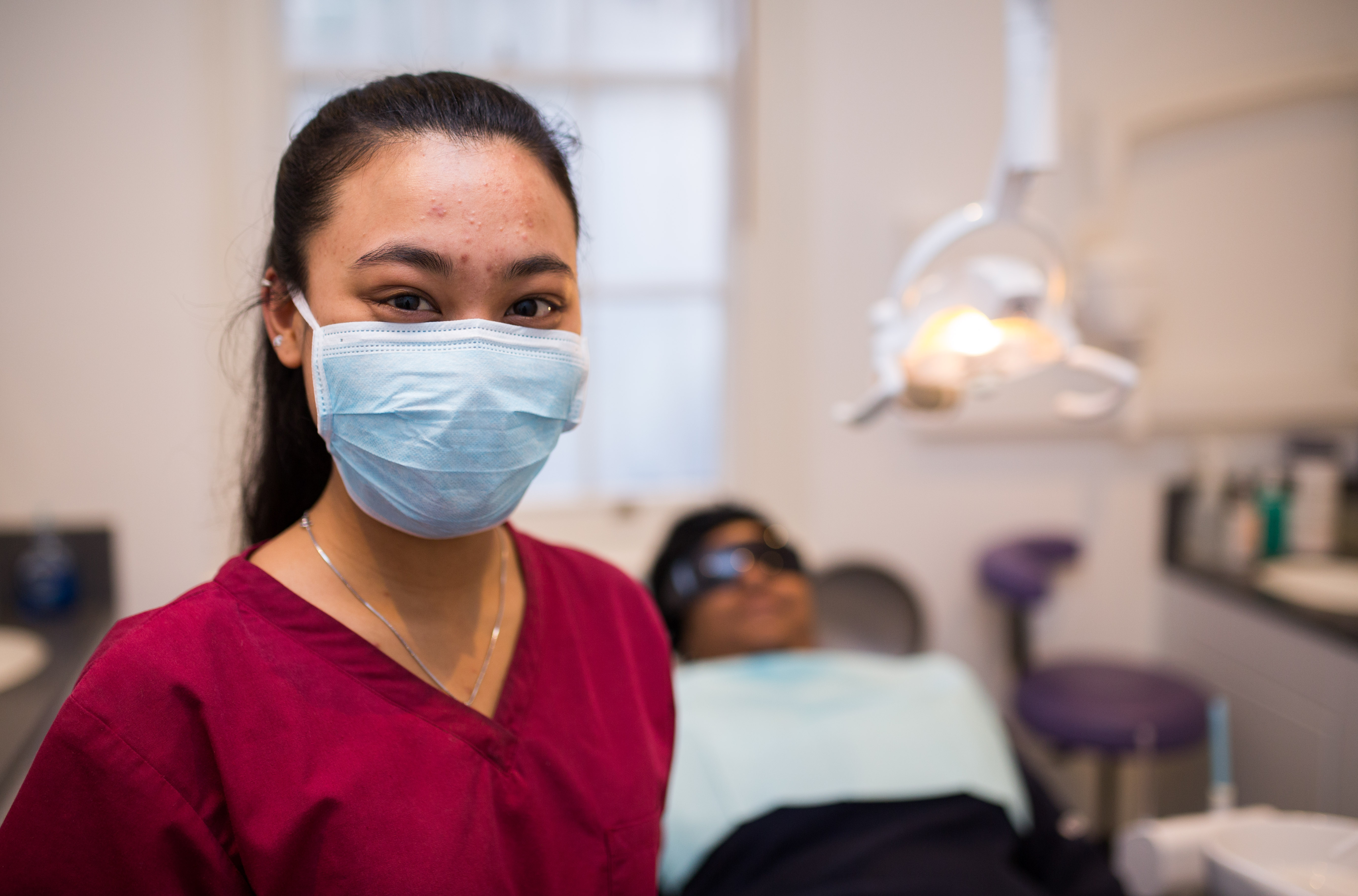 Dental nurse looks into camera, patient on dentists chair behind her 