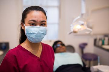 Dental nurse looks into camera, patient on dentists chair behind her 
