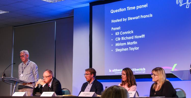 Our Question Time panel and host seated on a stage and listening to questions from the audience. Behind the panel is a large screen with their names on it.