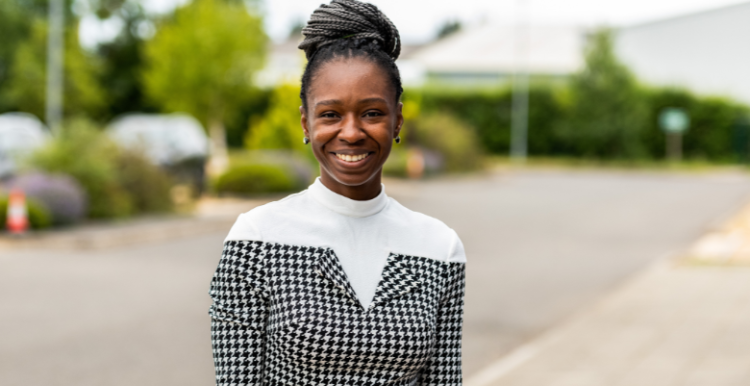 A woman standing in a street and smiling. There parked cars and bushes in the background