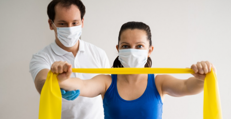 woman wearing a mask using a resistance band and a physio in a mask stands behind her