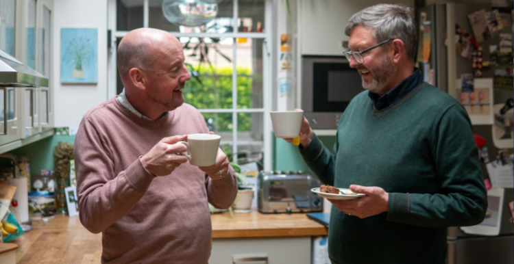 Two men standing in a kitchen having a cup of tea