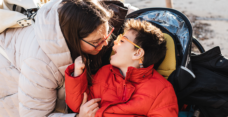 disabled boy in wheelchair outside wearing redcoat and woman leaning over to hug him