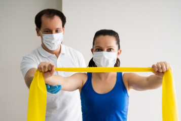 woman wearing a mask using a resistance band and a physio in a mask stands behind her